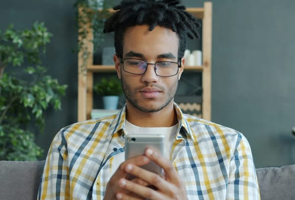 Happy Moddle Eastern man is playing video game at home using modern smartphone touching screen sitting at desk with laptop computer.